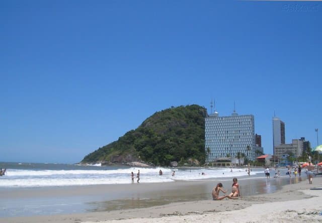 Banhistas aproveitando as ondas na orla de Caiobá com morro verde ao fundo