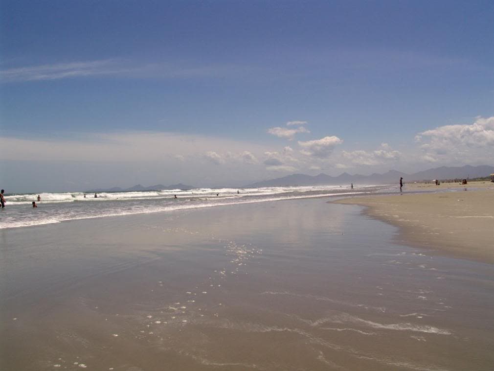 Balneário Shangri-lá com ampla faixa de areia molhada refletindo o céu e montanhas ao fundo