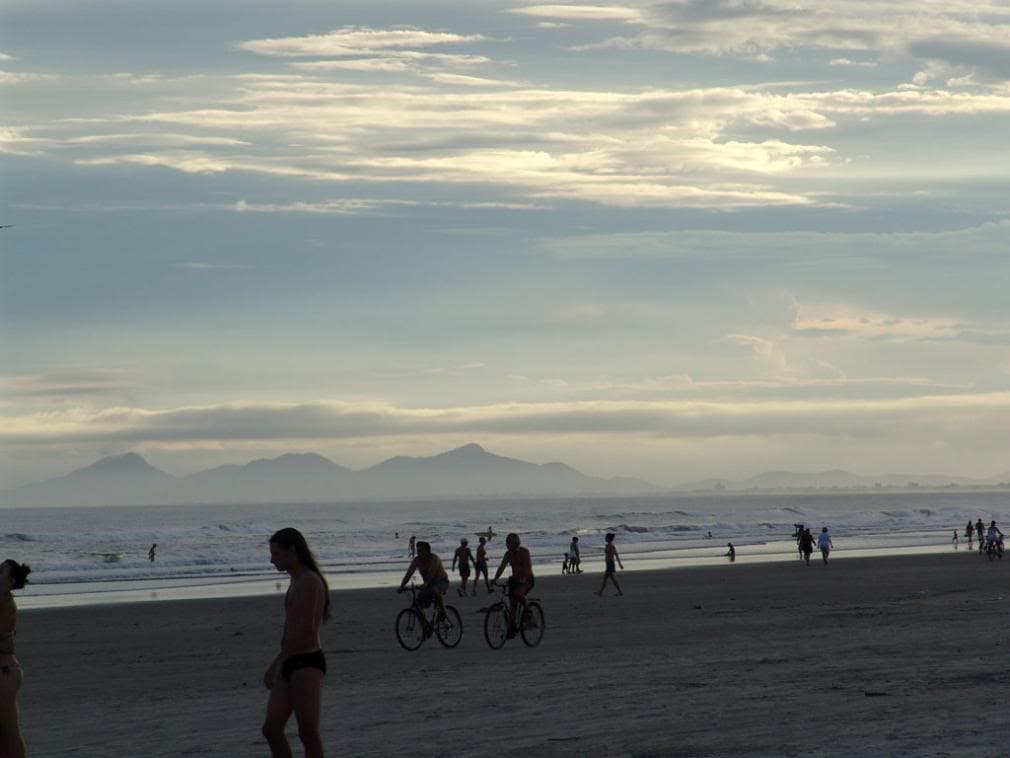 Faixa de areia escura com ondas e banhistas caminhando à beira-mar