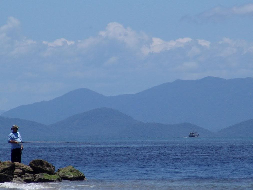 Costão rochoso com pessoa sobre pedras, mar e pequena embarcação ao fundo